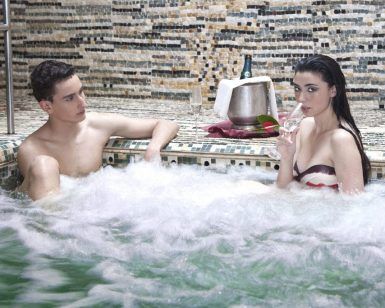 Dos personas se relajan en el jacuzzi de su patio trasero; una disfruta de una copa con una botella de vino en una nevera cercana, evocando el encanto de Sevilla. Es como vivir su propia experiencia del Circuito de Spa en las Termas de Hispalis en casa. - ForTwo - Planes para parejas