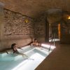 Dos personas se relajan en la piscina cubierta rústica y poco iluminada de Escapada con desayuno y spa en Balneario Caldes de Montbui (Barcelona), envuelta por paredes de piedra y techos abovedados. - ForTwo - Planes para parejas