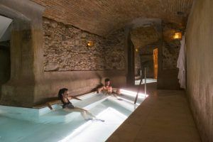 Dos personas se relajan en la piscina cubierta rústica y poco iluminada de Escapada con desayuno y spa en Balneario Caldes de Montbui (Barcelona), envuelta por paredes de piedra y techos abovedados. - ForTwo - Planes para parejas