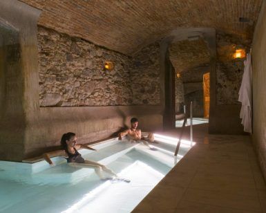 Dos personas se relajan en la piscina cubierta rústica y poco iluminada de Escapada con desayuno y spa en Balneario Caldes de Montbui (Barcelona), envuelta por paredes de piedra y techos abovedados. - ForTwo - Planes para parejas