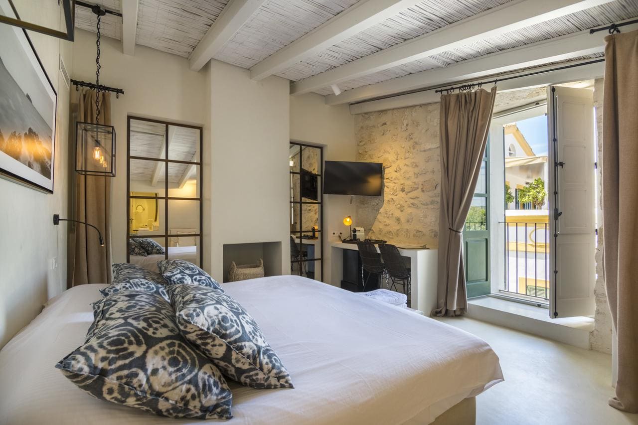 Disfrute de una acogedora habitación en el Hotel Torre del Canónigo (Ibiza), decorada con detalles en blanco, almohadas estampadas, televisor y acceso al balcón. Disfrute del encanto de las vigas de madera rústicas y los detalles de piedra en las paredes, creando un ambiente acogedor. - ForTwo - Planes para parejas