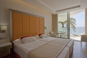 Disfrute de una habitación contemporánea en el Hotel Torre del Mar en Ibiza, equipada con dos camas individuales, un cabecero naranja y un balcón que ofrece impresionantes vistas al mar y palmeras ondulantes. - ForTwo - Planes para parejas