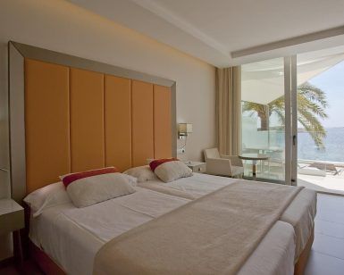Disfrute de una habitación contemporánea en el Hotel Torre del Mar en Ibiza, equipada con dos camas individuales, un cabecero naranja y un balcón que ofrece impresionantes vistas al mar y palmeras ondulantes. - ForTwo - Planes para parejas