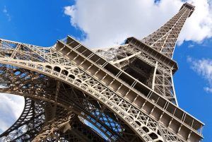 Vista en ángulo bajo de la Torre Eiffel contra un cielo azul nublado en París, mostrando su grandeza con un pase "Visita la Torre Eiffel sin hacer cola (París)". - ForTwo - Planes para parejas