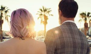 Una pareja, de espaldas, disfruta de un atardecer alicantino enmarcado por palmeras. La mujer de pelo rosa y el hombre con chaqueta a cuadros crean un momento perfecto en su "Sesión de fotos en pareja con Melina Bolopá (Alicante)". - ForTwo - Planes para parejas