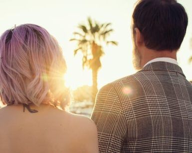 Una pareja, de espaldas, disfruta de un atardecer alicantino enmarcado por palmeras. La mujer de pelo rosa y el hombre con chaqueta a cuadros crean un momento perfecto en su "Sesión de fotos en pareja con Melina Bolopá (Alicante)". - ForTwo - Planes para parejas