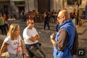 Tres personas charlan en una calle soleada, mientras otras están sentadas en mesas al aire libre al fondo, quizás comentando su reciente "Tour Guiado en Granada por Albaicín y Sacromonte" o planeando una aventura por el histórico barrio del Albaicín. - ForTwo - Planes para parejas