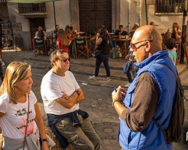 Tres personas charlan en una calle soleada, mientras otras están sentadas en mesas al aire libre al fondo, quizás comentando su reciente "Tour Guiado en Granada por Albaicín y Sacromonte" o planeando una aventura por el histórico barrio del Albaicín. - ForTwo - Planes para parejas