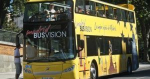 Un autobús amarillo brillante de dos pisos con la etiqueta BUSVISION recorre Madrid. El "Tour panorámico por Madrid en autobús de lujo" ofrece a los pasajeros vistas panorámicas desde el piso superior, haciendo que cada viaje sea inolvidable. - ForTwo - Planes para parejas