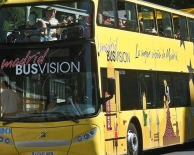 Un autobús amarillo brillante de dos pisos con la etiqueta BUSVISION recorre Madrid. El "Tour panorámico por Madrid en autobús de lujo" ofrece a los pasajeros vistas panorámicas desde el piso superior, haciendo que cada viaje sea inolvidable. - ForTwo - Planes para parejas