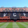 Las gradas del estadio exhiben grandes letras VCF con filas de asientos rojos y negros sobre el vibrante campo verde. Disfruta de esta icónica vista durante el tour Visita Mestalla con el Mestalla Forever en el famoso estadio del Valencia. - ForTwo - Planes para parejas