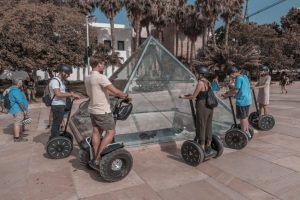 La gente que realiza la "Ruta en segway por Málaga" se desliza alrededor de una pirámide de cristal en un parque soleado con árboles al fondo. - ForTwo - Planes para parejas