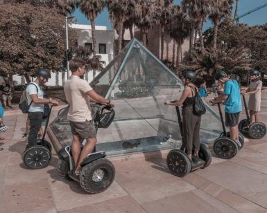 La gente que realiza la "Ruta en segway por Málaga" se desliza alrededor de una pirámide de cristal en un parque soleado con árboles al fondo. - ForTwo - Planes para parejas