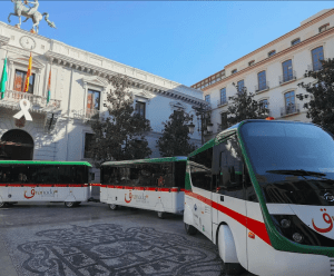 Tres autobuses eléctricos en verde y blanco, estacionados en una plaza con edificios históricos detrás, haciendo eco de las Entradas para Tren turístico en Granada, combinan la elegancia moderna con el encanto atemporal de Granada. - ForTwo - Planes para parejas