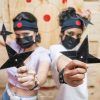 Dos personas, con diademas y máscaras, sostienen estrellas ninja enfocadas con un tablero de objetivos detrás de ellas, evocando el espíritu de Tiro con hacha en Madrid con El Hachazo, donde compiten los entusiastas del lanzamiento de hachas. - ForTwo - Planes para parejas