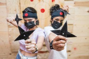 Dos personas, con diademas y máscaras, sostienen estrellas ninja enfocadas con un tablero de objetivos detrás de ellas, evocando el espíritu de Tiro con hacha en Madrid con El Hachazo, donde compiten los entusiastas del lanzamiento de hachas. - ForTwo - Planes para parejas