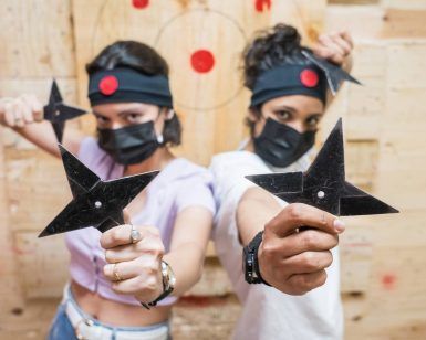 Dos personas, con diademas y máscaras, sostienen estrellas ninja enfocadas con un tablero de objetivos detrás de ellas, evocando el espíritu de Tiro con hacha en Madrid con El Hachazo, donde compiten los entusiastas del lanzamiento de hachas. - ForTwo - Planes para parejas