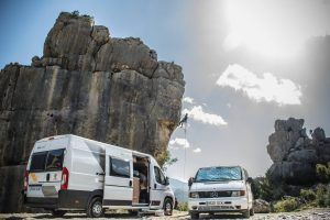 Dos Escapada romántica en furgoneta camperizada con Vantrip (Barcelona) están aparcadas junto a formaciones rocosas; un escalador escala la roca bajo un cielo soleado con nubes dispersas, ideal para este emocionante vantrip. - ForTwo - Planes para parejas