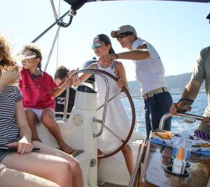La gente disfruta de un día soleado en una "Experiencia en velero en Barcelona + Vermú", con una persona al volante y otros relajándose en la cubierta, bebiendo Vermú. - ForTwo - Planes para parejas