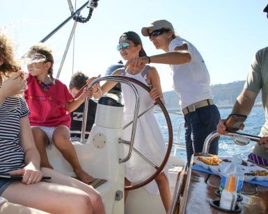La gente disfruta de un día soleado en una "Experiencia en velero en Barcelona + Vermú", con una persona al volante y otros relajándose en la cubierta, bebiendo Vermú. - ForTwo - Planes para parejas
