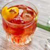 Un vaso de bebida helada oscura con un toque de naranja reposa elegantemente sobre una superficie de madera, rodeado de cubitos de hielo dispersos, evocando el encanto de una degustación de Cata de vermuts con aperitivo en Di-Ví en Barcelona. - ForTwo - Planes para parejas