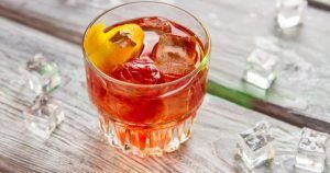 Un vaso de bebida helada oscura con un toque de naranja reposa elegantemente sobre una superficie de madera, rodeado de cubitos de hielo dispersos, evocando el encanto de una degustación de Cata de vermuts con aperitivo en Di-Ví en Barcelona. - ForTwo - Planes para parejas