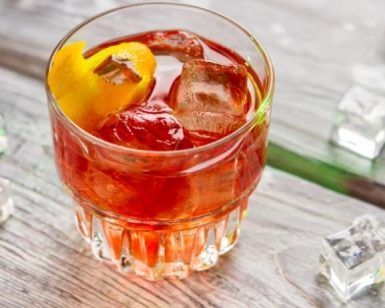 Un vaso de bebida helada oscura con un toque de naranja reposa elegantemente sobre una superficie de madera, rodeado de cubitos de hielo dispersos, evocando el encanto de una degustación de Cata de vermuts con aperitivo en Di-Ví en Barcelona. - ForTwo - Planes para parejas