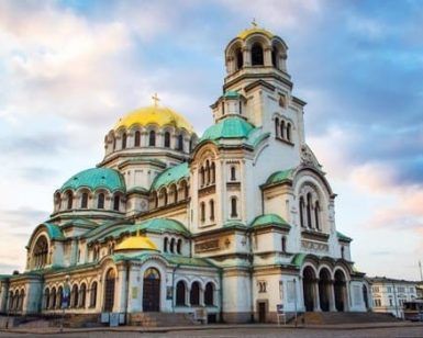 La pintoresca catedral, con su cúpula verde y dorada, se alza majestuosa contra un atardecer parcialmente nublado, capturando el patrimonio arquitectónico celebrado en "Descubre Rumanía y Bulgaria". - ForTwo - Planes para parejas