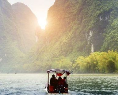 Un barco se desliza por un río con pasajeros bajo un cielo brumoso, enclavado entre altas y verdes montañas que recuerdan a la China clásica y a Hong Kong. - ForTwo - Planes para parejas