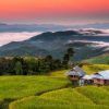 Un paisaje sereno al atardecer en Tailandia presenta una pequeña casa en medio de exuberantes campos verdes, colinas brumosas en la distancia y un cielo naranja, todos elementos de "Descubre los colores de Tailandia y Camboya". - ForTwo - Planes para parejas