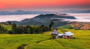 Un paisaje sereno al atardecer en Tailandia presenta una pequeña casa en medio de exuberantes campos verdes, colinas brumosas en la distancia y un cielo naranja, todos elementos de "Descubre los colores de Tailandia y Camboya". - ForTwo - Planes para parejas