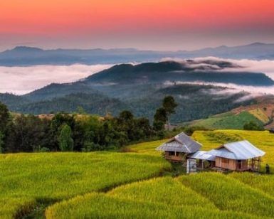 Un paisaje sereno al atardecer en Tailandia presenta una pequeña casa en medio de exuberantes campos verdes, colinas brumosas en la distancia y un cielo naranja, todos elementos de "Descubre los colores de Tailandia y Camboya". - ForTwo - Planes para parejas
