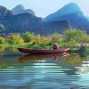 Un pequeño barco rojo en un río sereno en Vietnam, rodeado de exuberante vegetación y montañas bajo un cielo azul claro, crea una escena ideal durante el Viaje por Vietnam en 8 días. - ForTwo - Planes para parejas