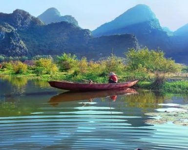 Un pequeño barco rojo en un río sereno en Vietnam, rodeado de exuberante vegetación y montañas bajo un cielo azul claro, crea una escena ideal durante el Viaje por Vietnam en 8 días. - ForTwo - Planes para parejas