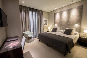 Alójese en una habitación contemporánea en el Hotel Vincci Mercat de Valencia, con cama doble, dos lámparas de noche, escritorio con silla, elegantes obras de arte en la pared y un amplio ventanal con cortinas. Disfrute de una experiencia excepcional llena de sofisticación. - ForTwo - Planes para parejas