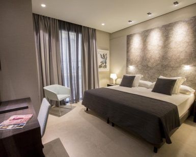 Alójese en una habitación contemporánea en el Hotel Vincci Mercat de Valencia, con cama doble, dos lámparas de noche, escritorio con silla, elegantes obras de arte en la pared y un amplio ventanal con cortinas. Disfrute de una experiencia excepcional llena de sofisticación. - ForTwo - Planes para parejas