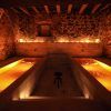 En una sala de spa poco iluminada inspirada en el Ritual Baño del Vino por Aire de Vallromanes - Barcelona, dos piscinas paralelas de agua tibia y resplandeciente invitan a la relajación frente a paredes de piedra rústica. - ForTwo - Planes para parejas