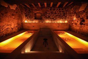En una sala de spa poco iluminada inspirada en el Ritual Baño del Vino por Aire de Vallromanes - Barcelona, dos piscinas paralelas de agua tibia y resplandeciente invitan a la relajación frente a paredes de piedra rústica. - ForTwo - Planes para parejas
