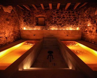 En una sala de spa poco iluminada inspirada en el Ritual Baño del Vino por Aire de Vallromanes - Barcelona, dos piscinas paralelas de agua tibia y resplandeciente invitan a la relajación frente a paredes de piedra rústica. - ForTwo - Planes para parejas