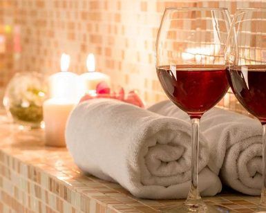 Dos copas de vino tinto y toallas enrolladas sobre una superficie de baldosas, con velas encendidas cerca, crean la magia de la vinoterapia. Disfrute de este capricho en Ambrosia Urban Spa en Barcelona. - ForTwo - Planes para parejas