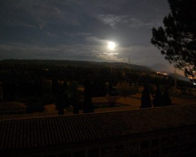 Un paisaje iluminado por la luna con árboles recortados, colinas y un tejado bajo un cielo nocturno nublado evoca el misterio de explorar el cementerio histórico de Granada durante la "Visita guiada privada al cementerio de Granada" bajo un dosel iluminado por las estrellas. - ForTwo - Planes para parejas