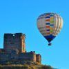 Un globo aerostático de "Un vuelo en globo en Madrid y alrededores" se desplaza con gracia cerca de las ruinas de un castillo de piedra bajo un cielo azul claro, ofreciendo impresionantes vistas de los alrededores. - ForTwo - Planes para parejas