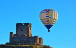 Un globo aerostático de "Un vuelo en globo en Madrid y alrededores" se desplaza con gracia cerca de las ruinas de un castillo de piedra bajo un cielo azul claro, ofreciendo impresionantes vistas de los alrededores. - ForTwo - Planes para parejas