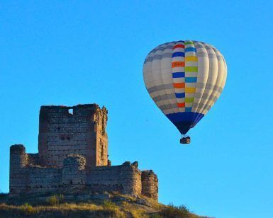 Un globo aerostático de "Un vuelo en globo en Madrid y alrededores" se desplaza con gracia cerca de las ruinas de un castillo de piedra bajo un cielo azul claro, ofreciendo impresionantes vistas de los alrededores. - ForTwo - Planes para parejas