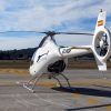 Un helicóptero blanco descansa elegantemente sobre un helipuerto con un paisaje montañoso como telón de fondo y un cielo parcialmente nublado, evocando paisajes serenos cerca del aeropuerto de Sabadell de Barcelona, perfecto para la experiencia "Vuelo en helicóptero desde el aeropuerto de Sabadell (Barcelona)". - ForTwo - Planes para parejas