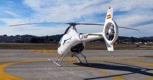 Un helicóptero blanco descansa elegantemente sobre un helipuerto con un paisaje montañoso como telón de fondo y un cielo parcialmente nublado, evocando paisajes serenos cerca del aeropuerto de Sabadell de Barcelona, perfecto para la experiencia "Vuelo en helicóptero desde el aeropuerto de Sabadell (Barcelona)". - ForTwo - Planes para parejas