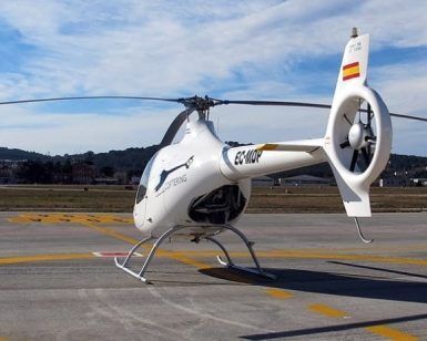 Un helicóptero blanco descansa elegantemente sobre un helipuerto con un paisaje montañoso como telón de fondo y un cielo parcialmente nublado, evocando paisajes serenos cerca del aeropuerto de Sabadell de Barcelona, perfecto para la experiencia "Vuelo en helicóptero desde el aeropuerto de Sabadell (Barcelona)". - ForTwo - Planes para parejas