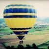 Un globo aerostático amarillo y azul se desplaza sobre un pintoresco paisaje rural con campos y árboles, evocando la tranquilidad de una experiencia de Vuelo en globo en Barcelona. - ForTwo - Planes para parejas