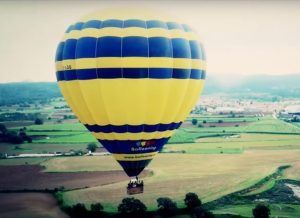 Un globo aerostático amarillo y azul se desplaza sobre un pintoresco paisaje rural con campos y árboles, evocando la tranquilidad de una experiencia de Vuelo en globo en Barcelona. - ForTwo - Planes para parejas
