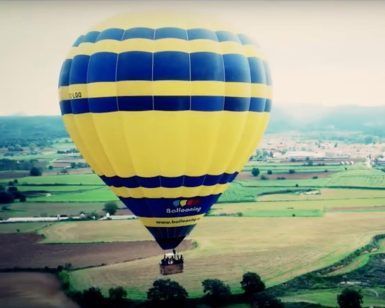 Un globo aerostático amarillo y azul se desplaza sobre un pintoresco paisaje rural con campos y árboles, evocando la tranquilidad de una experiencia de Vuelo en globo en Barcelona. - ForTwo - Planes para parejas